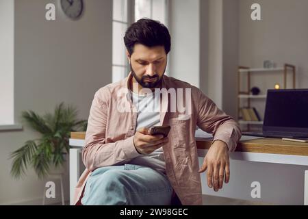 Bello e dai capelli scuri è seduto vicino al suo posto di lavoro sullo sfondo di un tavolo con un laptop. Foto Stock