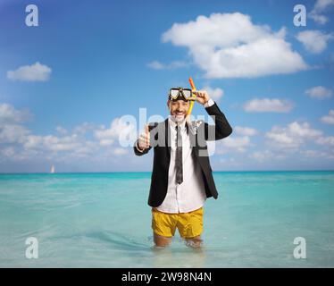 Uomo d'affari in piedi in mare con pantaloncini da nuoto gialli, maschera da immersione e pollici gestanti in alto Foto Stock