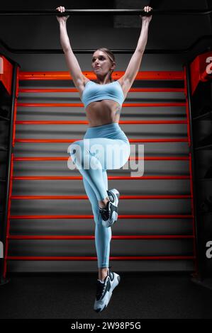 La bella bionda alta si tira sul bar della palestra sullo sfondo delle barre a muro. Il concetto di sport, fitness, aerobica, bodybuilding Foto Stock