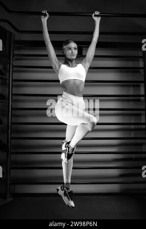 La bella bionda alta si tira sul bar della palestra sullo sfondo delle barre a muro. Il concetto di sport, fitness, aerobica, bodybuilding Foto Stock