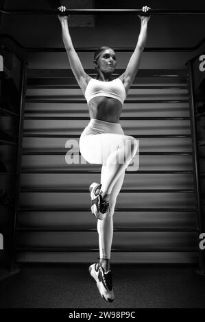 La bella bionda alta si tira sul bar della palestra sullo sfondo delle barre a muro. Il concetto di sport, fitness, aerobica, bodybuilding Foto Stock