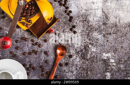 Vecchio macinacaffè con chicchi e caffè macinato accanto a una tazza vuota e a un cucchiaio di legno, su un tavolo in marmo grigio con più chicchi. Foto Stock