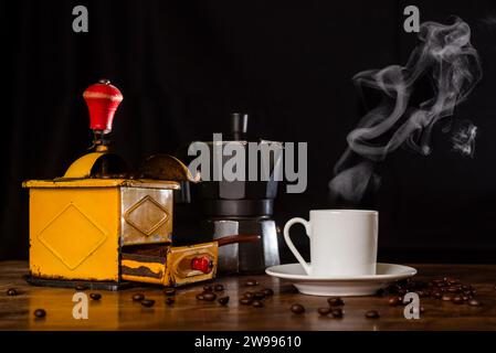 Vecchio macinacaffè accanto a una macchina per espresso e a una tazza di caffè fumante, vista frontale. Foto Stock