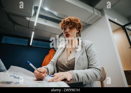 Adorabile donna d'affari che analizza statistiche diverse e scrive su carta mentre lavora da sola in ufficio. Foto Stock