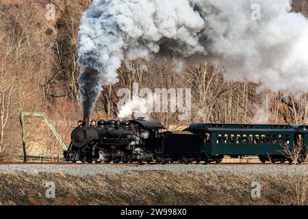 Vista di un treno passeggeri a vapore restaurato a scartamento ridotto, che soffia fumo nero e vapore bianco, viaggia attraverso le fattorie in un giorno d'inverno Foto Stock
