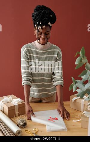 Felice ragazza afro-americana che avvolge il regalo di Natale in carta da imballaggio con stampa a mano di cervo rosso mentre si trova davanti alla macchina fotografica Foto Stock