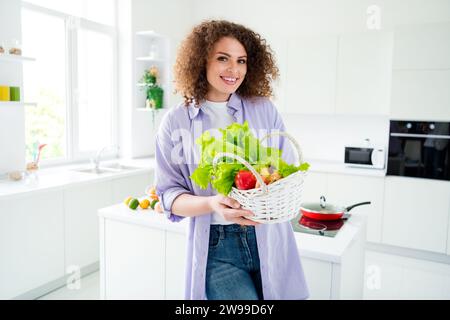 Foto di adorabile donna dolce che indossa una camicia viola che porta a casa verdure fresche del mercato all'interno della cucina dell'appartamento Foto Stock