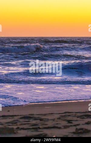 Un tramonto mozzafiato viene catturato su una spiaggia, con onde che si infrangono e un surfista solitario che cammina attraverso la sabbia Foto Stock