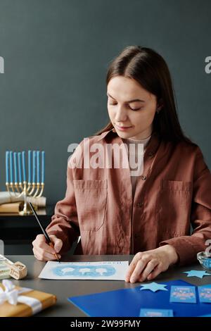 Giovane artigiana bruna in abbigliamento casual scrivendo i saluti su una cartolina Hanukkah fatta a mano con una fila di contorni dreidel Foto Stock