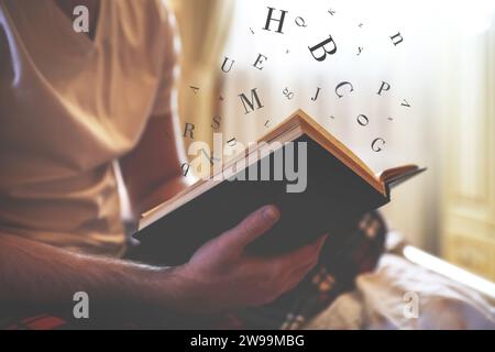 Giovane che legge un libro con lettere che volano sopra a letto a casa, primo piano Foto Stock