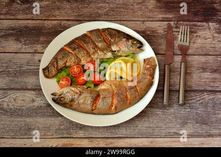 Piatto rotondo in ceramica con due trote intere al forno con lattuga, pomodori ciliegini e limone su un tavolo di legno. Vista dall'alto, base piatta. Foto Stock