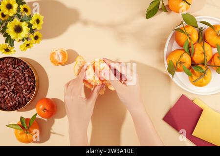 Le mani stanno pelando un mandarino, un vassoio di semi di melone essiccati, un piatto di mandarini, un bouquet di girasoli e buste portafortuna su uno schienale pastello Foto Stock