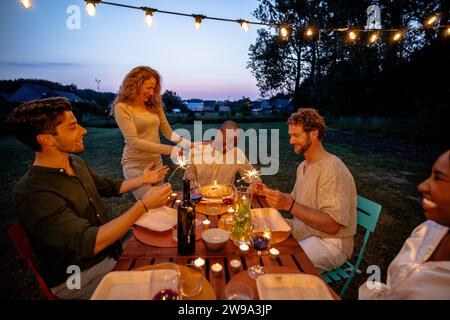 Al calar del tramonto, un'elegante cena all'aperto è in pieno svolgimento. Una donna caucasica si alza per illuminare uno sparkler, aggiungendo magia al momento, mentre un uomo nero seduto al tavolo condivide una risata con lei. Un uomo caucasico e una donna di origine africana, entrambi seduti, si uniscono alla celebrazione. Il tavolo è allestito splendidamente con bicchieri da vino e candele sotto un baldacchino di luci da favola, creando un'atmosfera affascinante al crepuscolo. Serata di eleganza: Cena all'aperto sotto le Fairy Lights. Foto di alta qualità Foto Stock