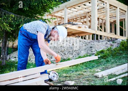 Falegname che costruisce casa in legno-incorniciata. L'uomo misura le distanze utilizzando un metro a nastro mentre indossa abiti da lavoro e casco. L'idea alla base di una costruzione ecologicamente sana e contemporanea. Foto Stock