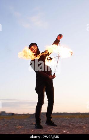 Ritratto verticale a tutta lunghezza di un uomo irriconoscibile che gira il fuoco durante lo spettacolo di fuoco al tramonto Foto Stock