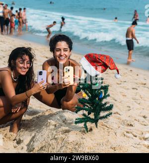Rio de Janeiro, Brasile - 25 dicembre 2023: Le giovani donne scattano foto con i loro smartphone di un albero di Natale in miniatura piantato sulla sabbia Foto Stock