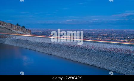 Fai il crepuscolo su una diga nel lago artificiale di Horsetooth, ai piedi delle Montagne Rocciose, in un paesaggio al crepuscolo autunnale o invernale con luci di traffico autostradale e distand Foto Stock