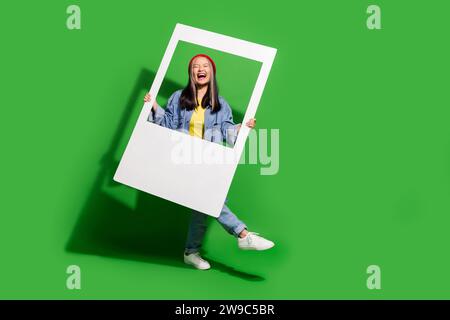 Foto del corpo di una giovane donna attraente che fa la danza della finestra indossa abiti di tendenza in jeans isolati su sfondo di colore verde Foto Stock