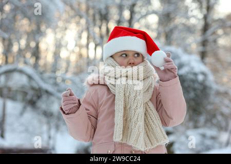 Graziosa bambina che indossa il cappello di Babbo Natale nel parco innevato il giorno d'inverno Foto Stock