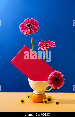 Gerberas fresche e buste rosse sono collocate in una tazza da tè in ceramica, circondata da piccoli fiori gialli. Sfondo blu beige. Spazio copia con la via anteriore Foto Stock