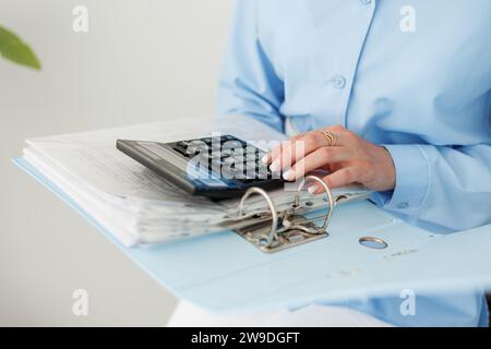 Foto ritagliata di una contabile in camicia blu calcola i costi della calcolatrice, conta i soldi, il budget aziendale, analizza i report finanziari, controlla la fattura Foto Stock