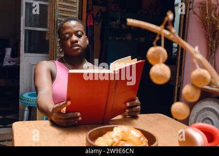 Giovane donna haitiana con i capelli corti, lettura tranquilla a casa seduta e tenendo un libro con le mani tese sul tavolo, concetto di stile di vita, spazio per la copia. Foto Stock
