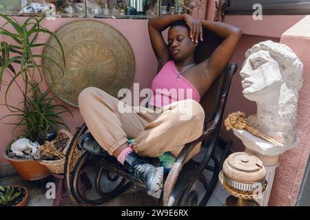 giovane donna haitiana dai capelli corti che riposa con gli occhi seduti e le braccia alzate, le gambe incrociate sono sedute su una sedia a dondolo di legno a casa Foto Stock