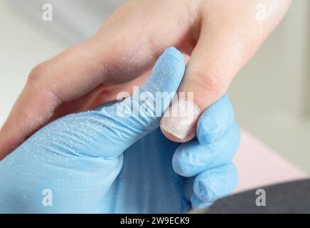 Processo di manicure nel salone di bellezza. Manicurista in guanti blu tiene il dito con un'unghia lucida. Foto Stock