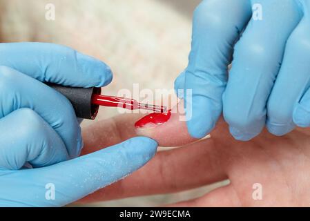 Manicurista in guanti blu che ricoprono l'unghia con smalto gel rosso. Processo di manicure in un salone di bellezza da vicino. Foto Stock