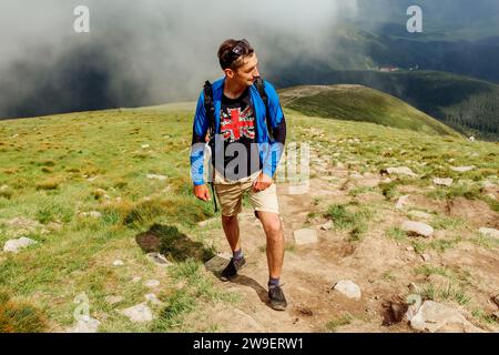 Uomo sportivo escursionista arrampicata su montagna circondata da nuvole in Carpazi. Stanco viaggiatore con zaino riposo. Viaggio in estate Ucraina Foto Stock