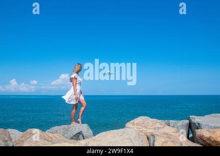 Beyond the Shore: Girl in White Dress, Stones, Sea, and an Airborne Plane Foto Stock