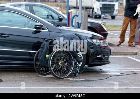 Riga, Lettonia, 4 gennaio 2021: Il conducente handicappato su sedia a rotelle pompa gli pneumatici della sua auto dal compressore in una stazione di benzina Foto Stock