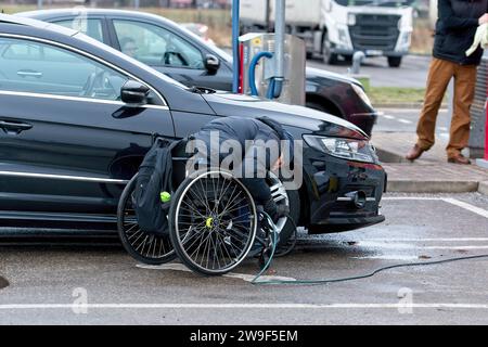 Riga, Lettonia, 4 gennaio 2021: Il conducente handicappato su sedia a rotelle pompa gli pneumatici della sua auto dal compressore in una stazione di benzina Foto Stock