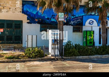 Una stazione di ricarica per veicoli elettrici si trova in un parcheggio a Marsaskala, Malta, davanti a una facciata dipinta con un murale a tema subacqueo che reca lo slogan “stesso pianeta, mondo diverso”. Foto Stock
