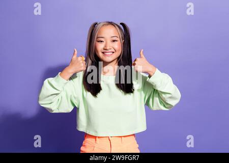 Ritratto di una ragazza asiatica soddisfatta con trecce di cavallo indossa un pullover oversize con due braccia che mostrano i pollici in alto isolati su sfondo di colore viola Foto Stock