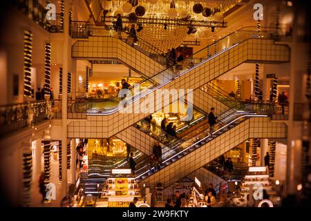 Grandi magazzini le Bon Marché, Parigi, Francia | grandi magazzini affollati con scale mobili multiple e illuminazione decorata, catturati in toni caldi. Foto Stock