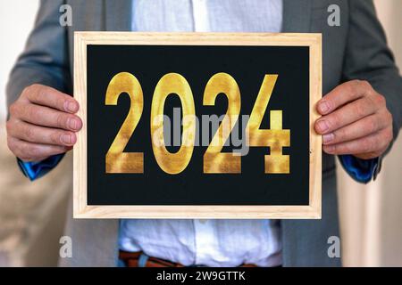 28 dicembre 2023: Uomo in costume che regge una targa con il numero 2024 in lettere d'oro. FOTOMONTAGGIO concept Capodanno e Capodanno *** Mann in Anzug hält eine Tafel mit der Zahl 2024 in goldener Schrift. Neujahr und Silvester Konzept FOTOMONTAGE Foto Stock