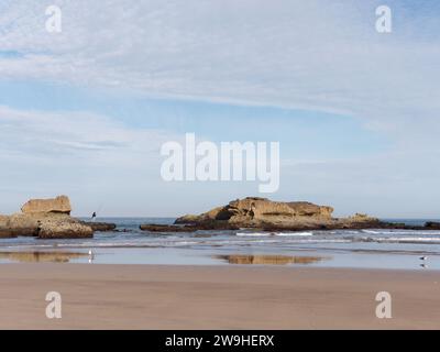 L'uomo pesca in cima a un affioramento roccioso sul bordo di una spiaggia sabbiosa nella città di Essaouira, in Marocco. 28 dicembre 2023 Foto Stock