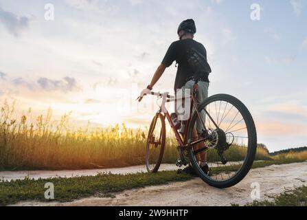 Ciclista con casco in piedi sul campo e godersi il tramonto dopo l'allenamento sportivo all'aria aperta. Attività all'aperto. Foto Stock