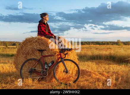Il giovane si siede su un pagliaio con una bici in un campo all'alba. Ciclista in un campo che si gode il tramonto. Concetto di stile di vita attivo. Foto Stock