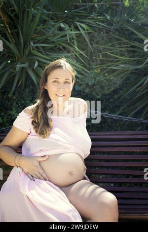 Donna incinta di sette mesi seduta su una panca di legno. Espressione molto felice Foto Stock