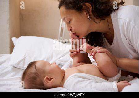 L'adorabile bambino di quattro mesi sorride guardando sua madre, mentre bacia i suoi piedi e piedi piccoli, godendosi momenti felici insieme. Delizioso Foto Stock