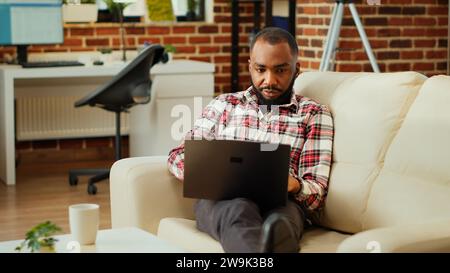 Semplice dipendente afroamericano che lavora da casa con un computer portatile, Teleworker che svolge varie attività in remoto, digitando sulla tastiera in un elegante e accogliente ufficio personale Foto Stock