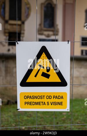 Segnale di avvertimento (triangolo giallo) su una recinzione intorno al cantiere (in un cimitero): Pericolo di caduta di oggetti dall'alto (testo in lingua croata) Foto Stock