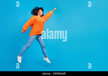 Foto a grandezza naturale di una graziosa ragazza che salta da supereroe che vola vestito da elegante abito a maglia arancione isolato su sfondo blu Foto Stock