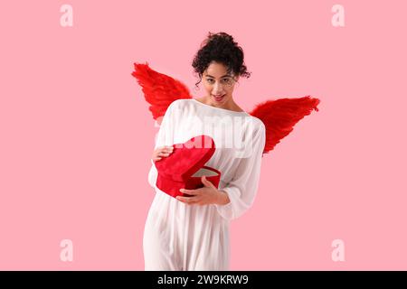 Giovane donna afro-americana vestita da Cupido che apre la confezione regalo su sfondo rosa. Festa di San Valentino Foto Stock