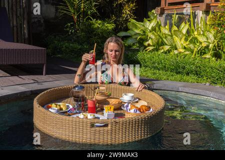 Una giovane donna che fa colazione su un cesto galleggiante in una piscina, Bali rurale Foto Stock