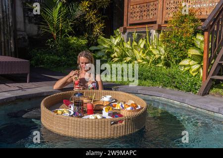 Una giovane donna che fa colazione su un cesto galleggiante in una piscina, Bali rurale Foto Stock