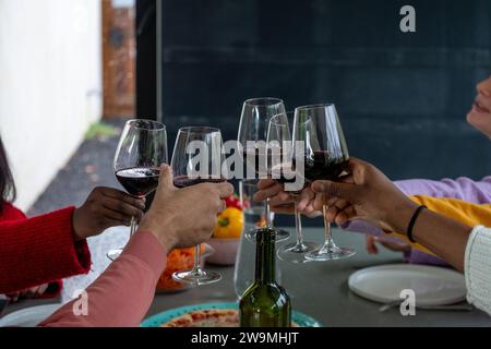 Un gruppo di persone si è riunito intorno a un tavolo da pranzo, per un brindisi gioioso con bicchieri di vino rosso. L'attenzione è rivolta alle mani e ai bicchieri da vino, per catturare un momento di festa e di cameratismo. L'ambiente suggerisce un intimo ritrovo interno, con una pizza parzialmente visibile e frutta sul tavolo, a indicare un pasto informale. Lo sfondo scuro contrasta con i colori vivaci indossati dagli individui, enfatizzando il calore dell'occasione. Brindisi celebrativi tra amici. Foto di alta qualità Foto Stock