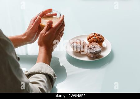 Primo piano di mani che reggono una tazza di caffè di vetro con latte e tre muffin di cagliata sul piatto sul lato destro. Snack delizioso e aromatico Foto Stock